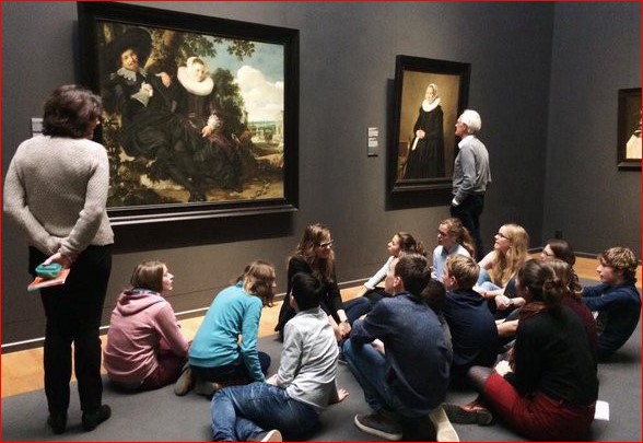 Un gruppo di ragazzi seduti per terra osserva attentamente un dipinto all'interno di un museo. Davanti a loro, una persona in piedi tiene un libro in mano, probabilmente una guida o un insegnante, mentre un uomo più anziano si trova di fronte a un altro quadro. Le opere d'arte esposte sembrano essere ritratti storici, e l'atmosfera nella sala è tranquilla e riflessiva, tipica di una visita guidata o di una lezione in un contesto educativo.